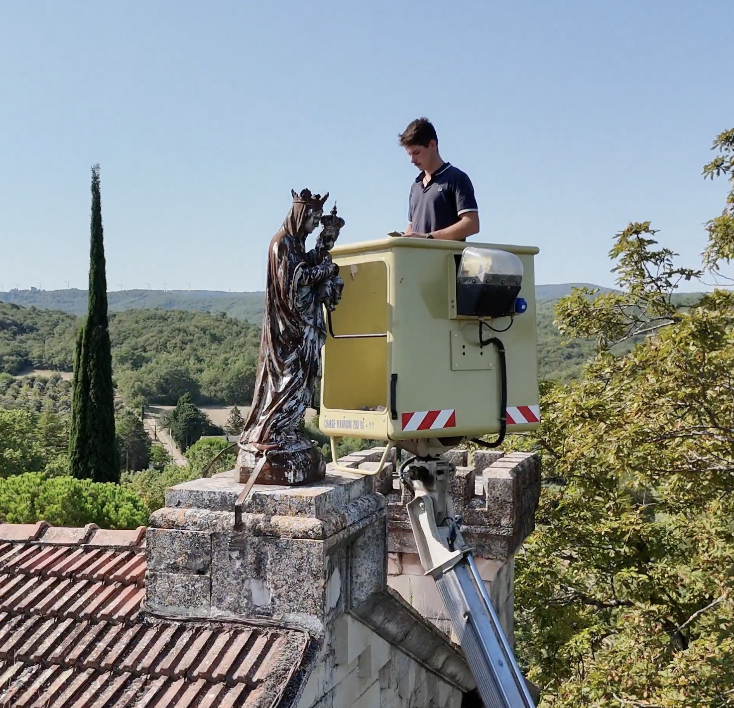 Raphaël Castex working beside a statue of Mary and the Child Jesus above the rooftops of Puygiron, France